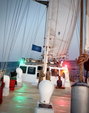 Front deck of Infinity ship while sailing at sea. They are navigation lights, Earth Flag and person sitting in cross legged meditation position by the deck house.