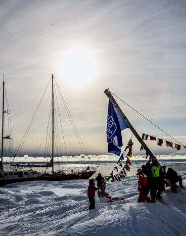 Earth Flag is being erected by Infinity expedition crew in the High Arctics - North West Passage. Snow covered landscape surrounds the scene.